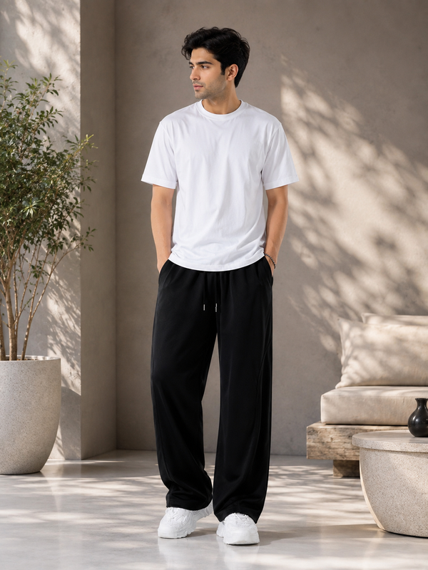 A man wearing a plain white short-sleeve crewneck t-shirt and black drawstring sweatpants, standing in a modern, sunlit indoor space with a concrete wall and potted plants.