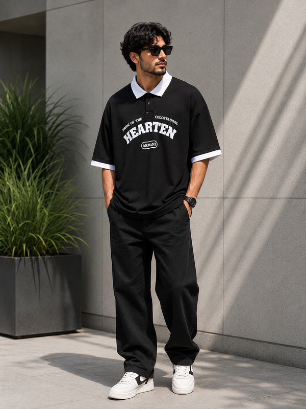 A man wearing a black short-sleeve polo shirt with white trim and a white 'HEARTEN' logo on the front, paired with black pants and white sneakers, standing against a gray concrete wall.