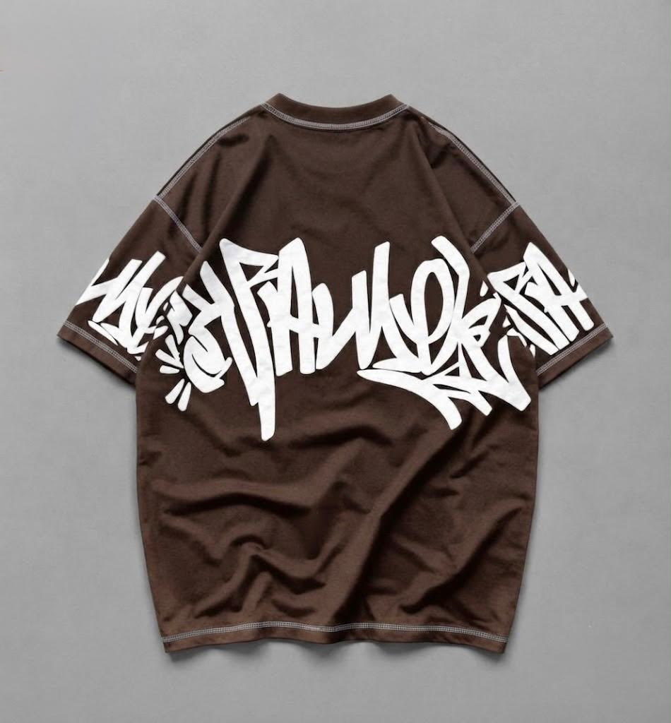 A brown short-sleeve t-shirt with a large white graffiti-style graphic printed across the front, displayed flat against a neutral gray background.